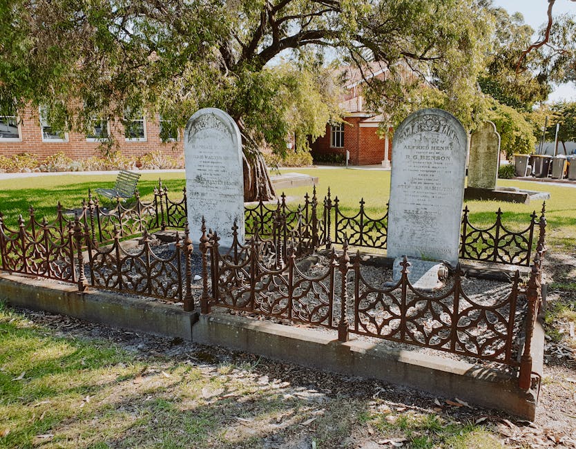 A small graveyard with two upright marble headstones surrounded by an ornate, rust-colored wrought iron fence with decorative finials. The headstones are weathered, with inscriptions partially visible