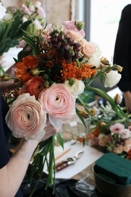 A close-up of a hand holding a large, fresh bouquet of mixed flowers, including soft pink ranunculus, pale peach roses, deep purple lisianthus, and vibrant orange blooms, arranged with lush green foli