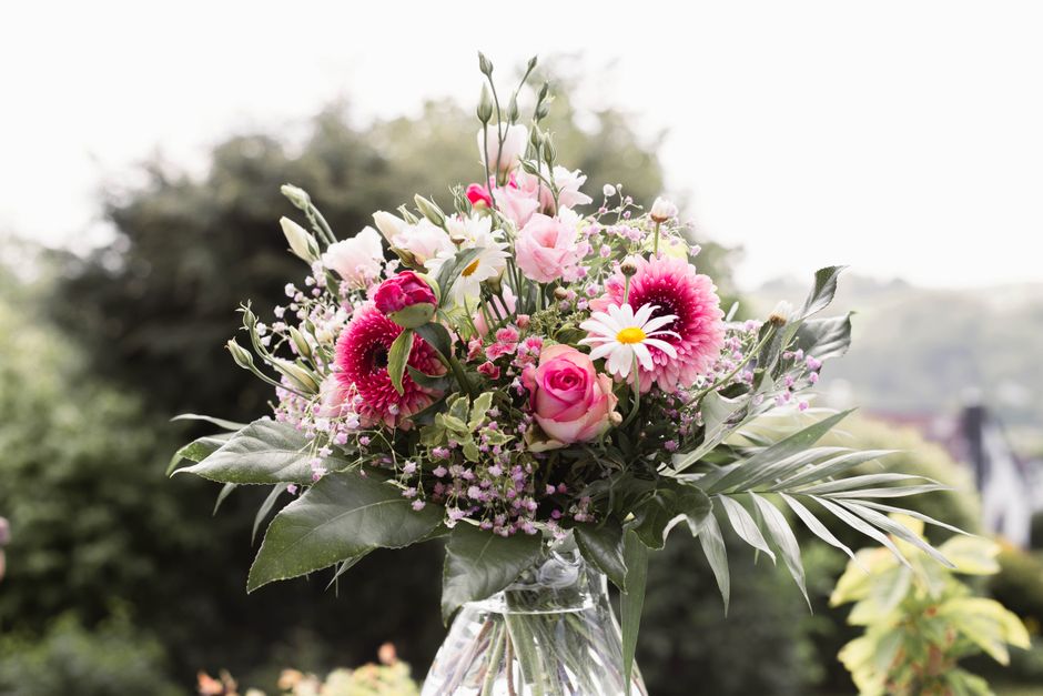 A fresh floral bouquet featuring a mix of pink and white flowers, including roses, daisies, and gerbera daisies, arranged with lush green foliage such as large leaves and smaller sprigs in a clear gla