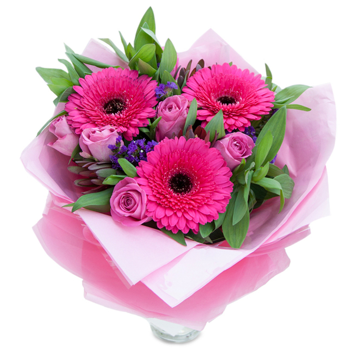 I imagine placing this bouquet on a small round table under soft morning light, its colour story led by three vivid fuchsia Gerbera daisies that open like three confident suns. Each Gerbera's many petals curl at the tips and draw the eye toward dark, ink-like centers that anchor the arrangement visually. Softer in temperament are the lavender-pink roses tucked in between - their petals layered in compact, spiralling folds that suggest freshness and restraint, a tender counterpoint to the daisies' exuberance. Flecks of purple statice are woven through the massed blooms; their tiny, clustered florets add a papery texture and a cooling tonal shift that creates depth without overpowering. A generous framework of green foliage - broad, slightly glossy leaves that read as salal or eucalyptus - cradles the flowers and contributes a crisp, vegetal scent when the bouquet is handled, reinforcing the sense of garden freshness. The florist has wrapped the stems with an inner sheet of soft matte pale pink tissue for a gentle base, then layered a slightly crinkled translucent fuchsia-pink cellophane for a subtle shimmer and a cohesive finish; the wrapping is folded with deliberate care, revealing the top rim of a clear glass vase below and promising immediate display. The overall feeling is celebratory yet intimate - a floral embrace suited to birthdays, Mother's Day, anniversaries, or as a thoughtful pick-me-up - reflecting the craft and attention to detail you'd expect from a local florist serving Brunswick Park's community.